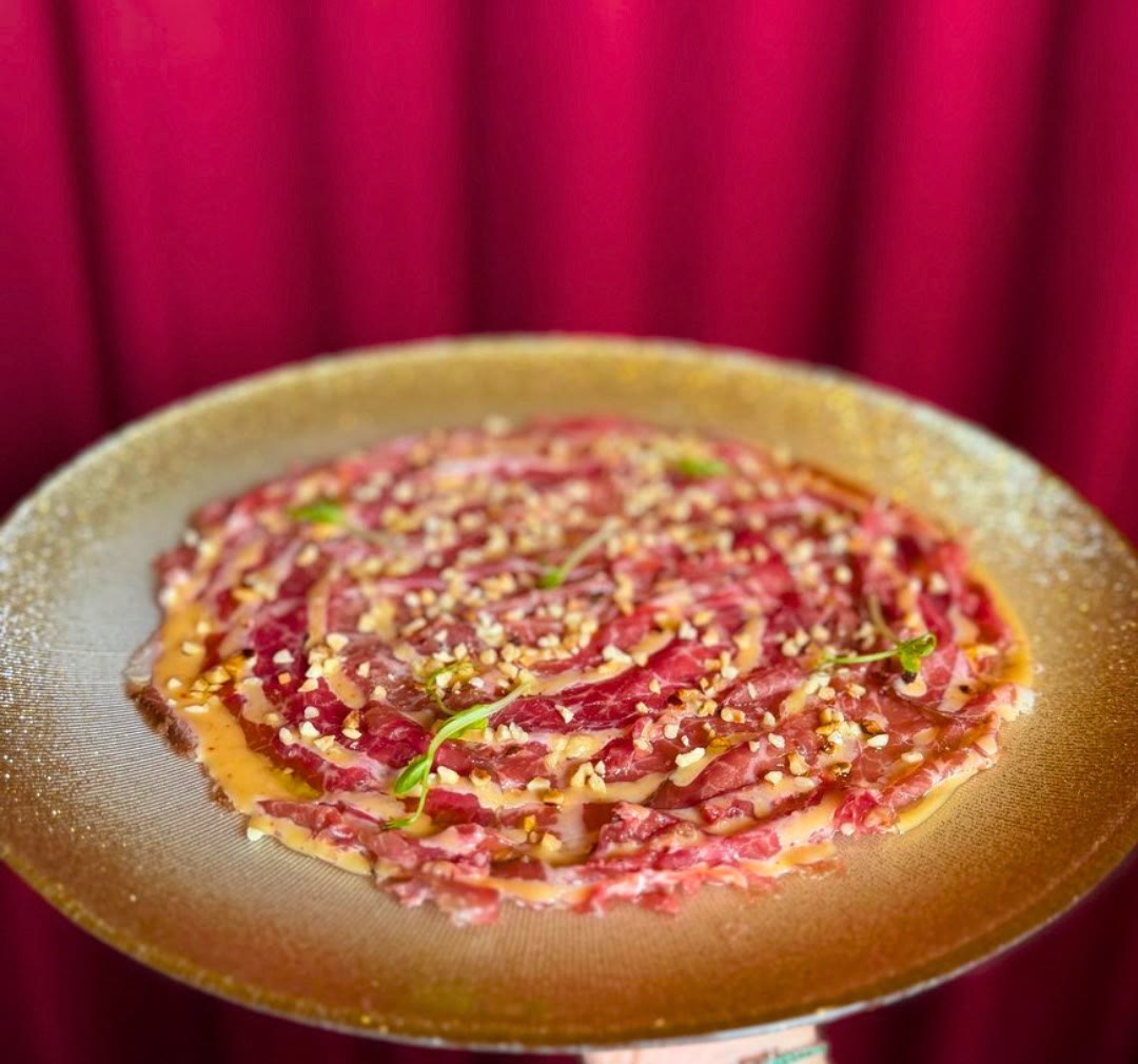 Carpaccio di carne con granella su un piatto dorato, davanti al velluto rosso